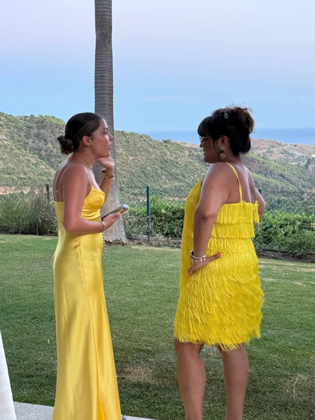 Nishma and Anushka in yellow dresses, deep in conversation on a hillside at sunset